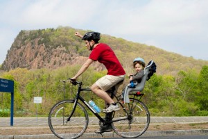 A young man rides a bike carrying a young child in a seat. He pumps his fist and cheers during the Rock to Rock Earth Day ride.