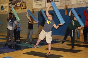 Rows of students stand on yoga mats and hold foam rollers over their heads during a class in flexibility at the JCC.