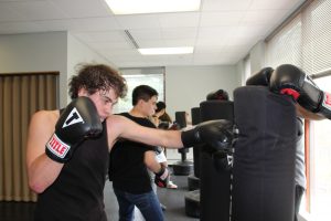 A high school student wearing boxing gloves works out during a strike class by hitting a punching bag.